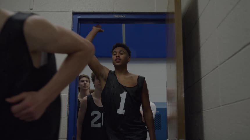 Several young male basketball players wearing dark numbered jerseys and shorts quickly exit a room through a blueframed doorway with one older man in a white shirt seen standing and clapping in the background