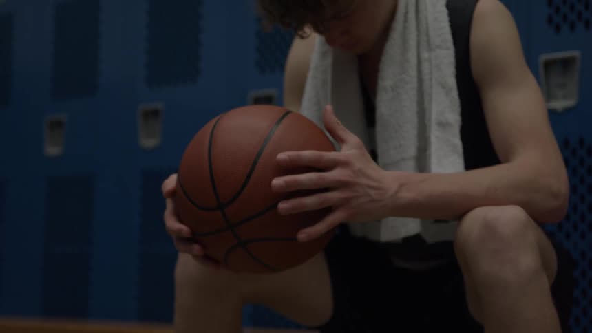 A young male basketball player sits with his head down on a wooden bench holding a brown basketball with both hands with a white towel draped around his neck and a wall of dark blue metal lockers visible behind him