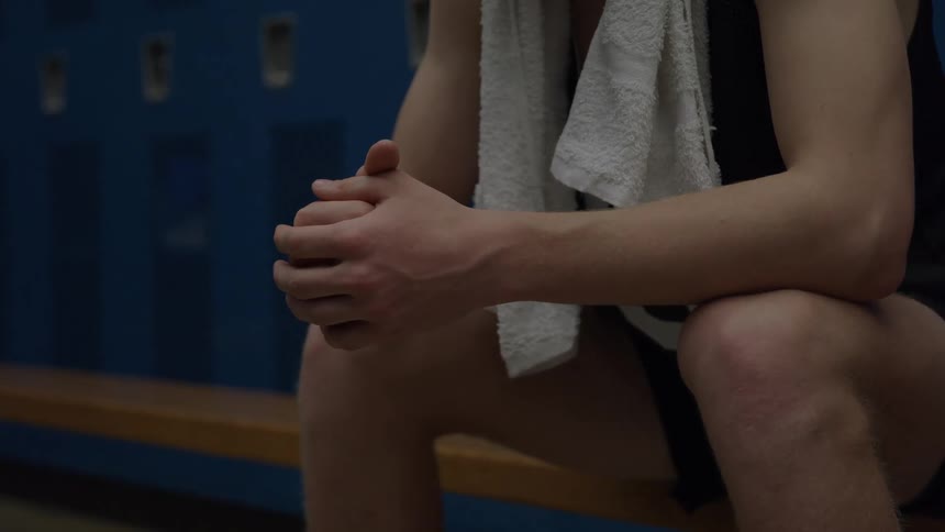 A close lower body view of a male basketball player sitting on a wooden bench with clasped hands and a white towel around his neck in a dimly lit blue locker room