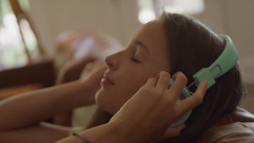A close profile view shows a teenage girl with long brown hair placing a pair of mint green and pink headphones over her ears before closing her eyes to relax on a brown sofa in a sunlit room