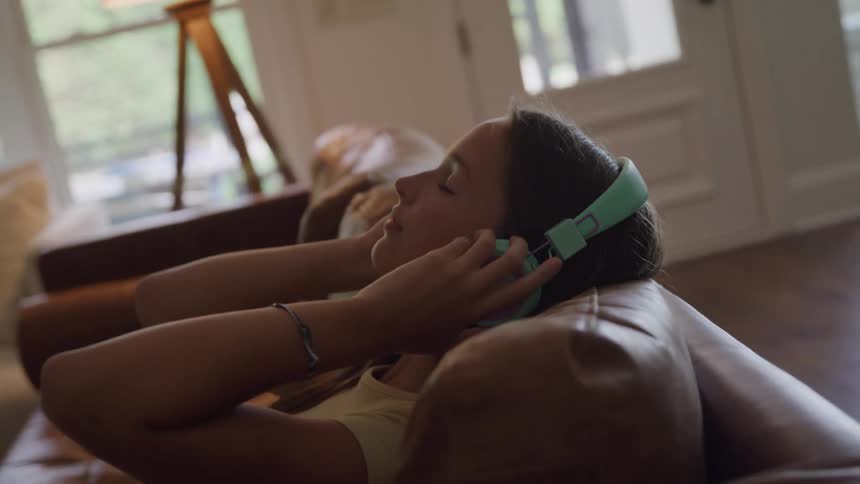 A young girl puts on colorful headphones and relaxes on a leather couch in a sunlit living room