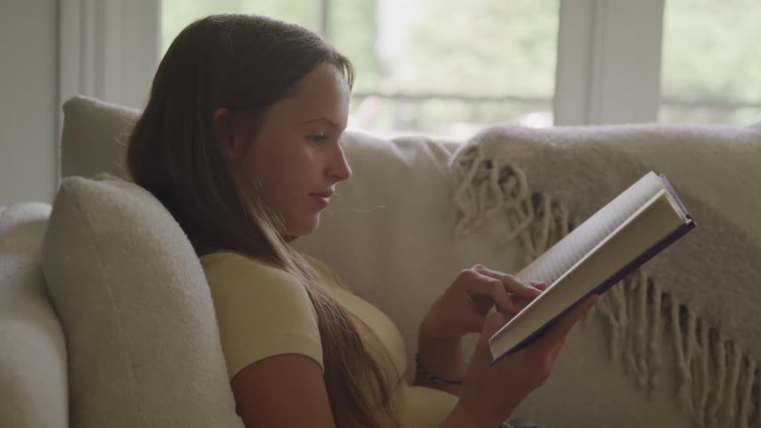 A medium profile view shows a young woman with long hair wearing a yellow shirt sitting comfortably on a white sofa silently reading a purple covered book with her finger following the words