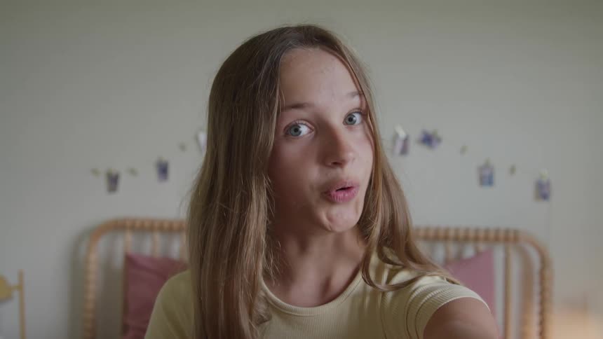 A teen girl smiles and waves while facing forward in a bedroom