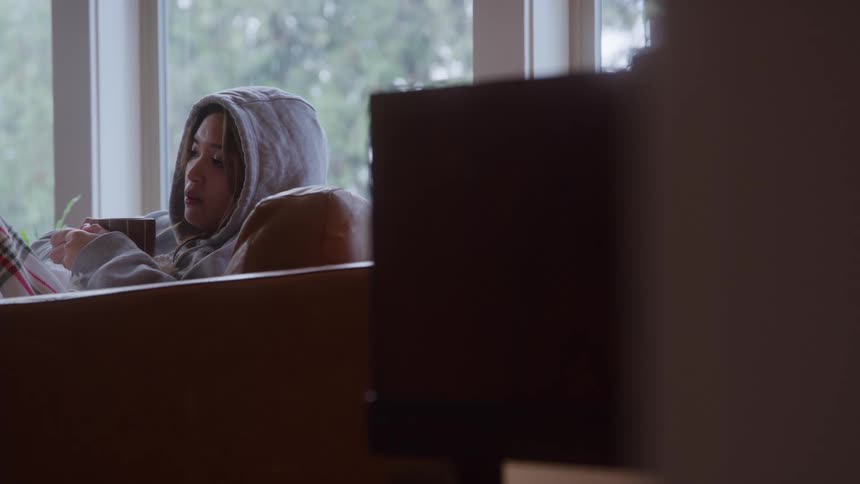 A woman in a hoodie sips from a striped mug while reading on a couch beside a large window on a gray day