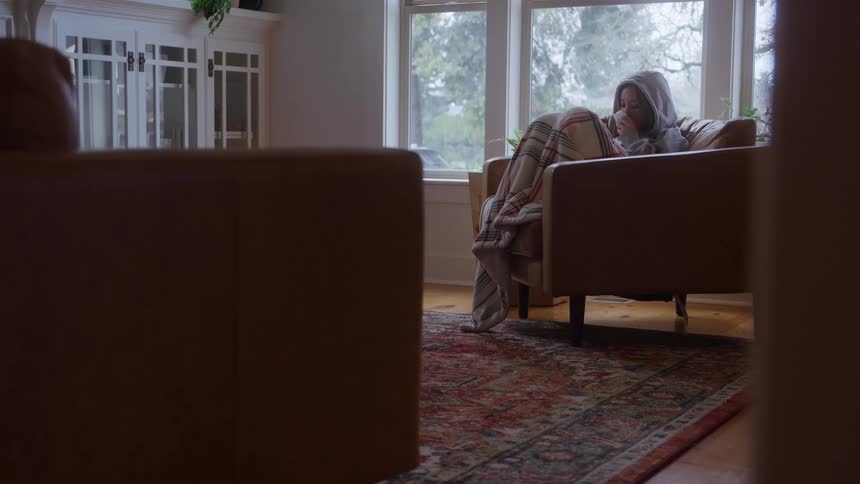 A young woman sits in an armchair in the living room wrapped in a hoodie and covering her feet with a blanket to stay warm sipping a cup of coffee in cozy comfort