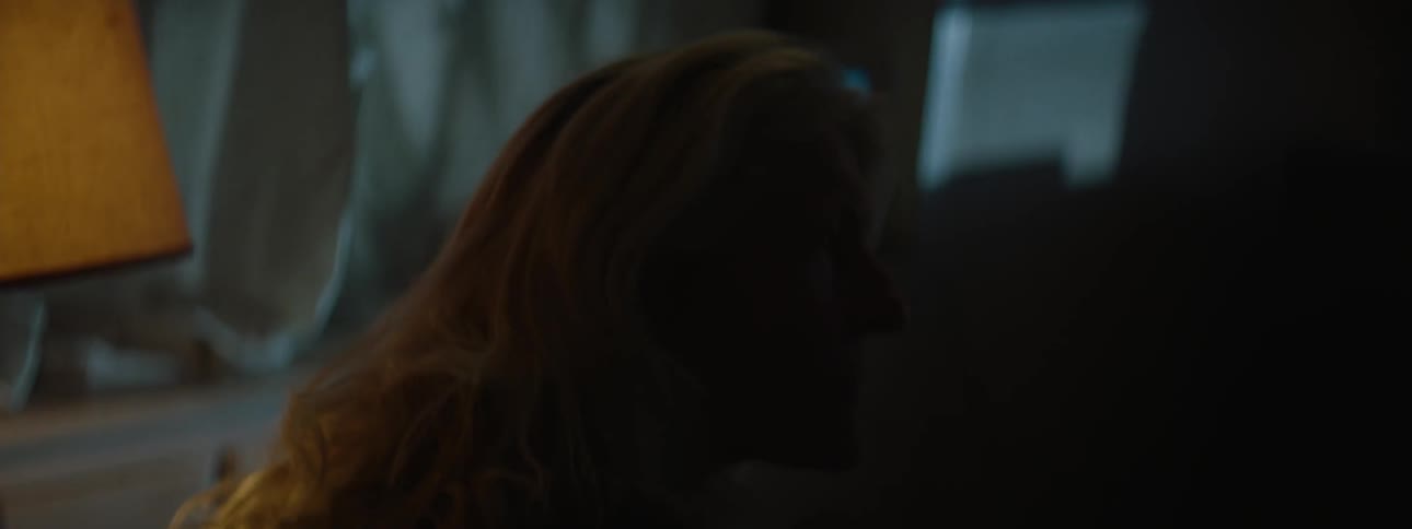 A woman sits in a dark living room as lights start to shine in the window