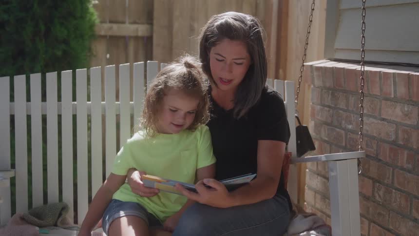 A woman and a young child sit together on a white porch swing outdoors with the woman holding and reading aloud from a book to the child while a white wooden fence and a red brick wall are visible in the background