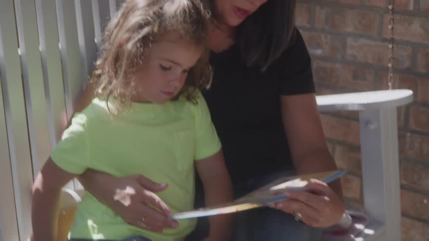 A woman sits on a porch step reading a picture book with a young girl in a neon green shirt who looks focused