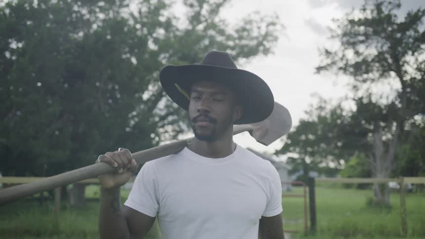 A strong man in a cowboy hat walks purposefully across a field carrying a shovel over his shoulder in a rural hazy setting