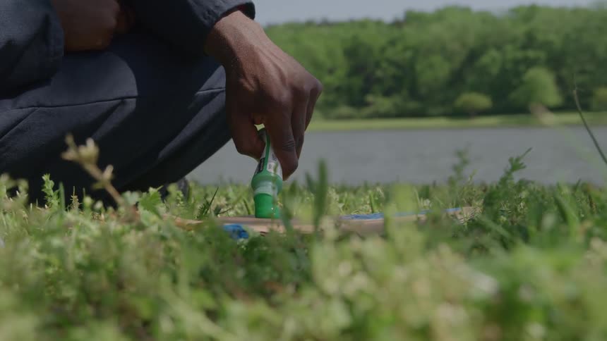 A closeup view of long green grass in the foreground shows a person squatting on the ground holding a small green plastic bottle and squeezing paint onto an object on the ground with a body of water and a line of trees visible in the distance