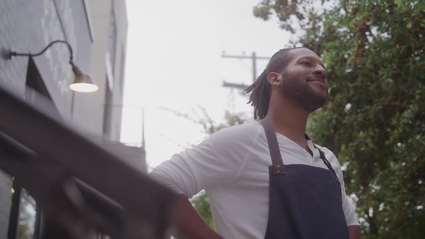 A man wearing a white shirt and dark apron stands outside with hands on his hips looking pleased beside a storefront lamp and leafy trees