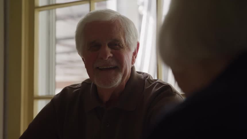 A closeup shot of an elderly man with white hair sitting at a table smiles and looks at another person whose shoulder and head are unfocused in the foreground with bright daylight entering the room through a large window