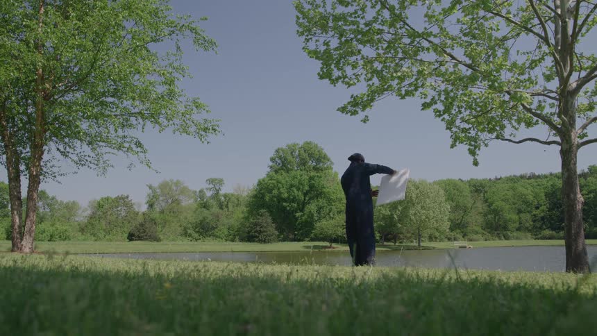 The video is a stationary wide shot of a lush green park scene with a pond or lake surrounded by trees under a bright blue sky in the latter half of the clip a man in dark clothing and a hat walks from the left across the foreground grass c