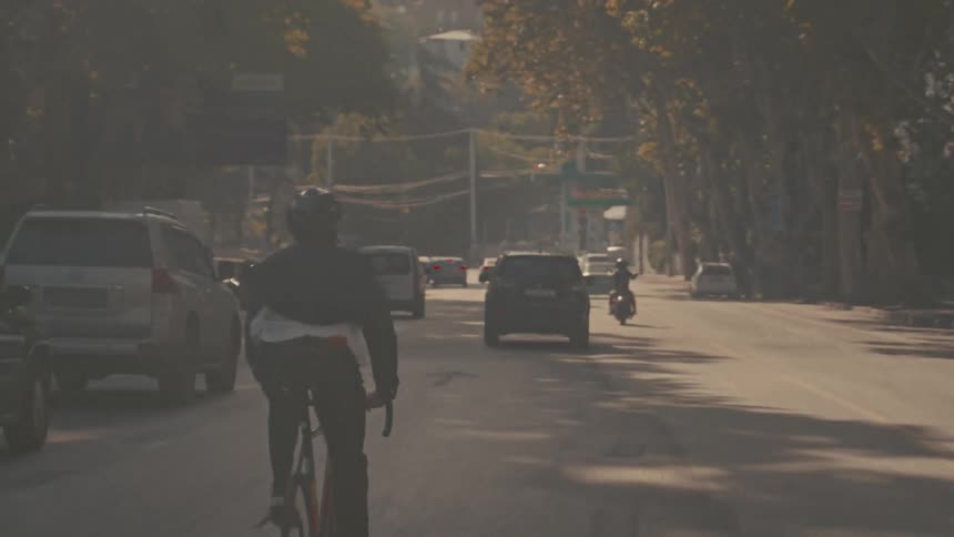 The video is a wide shot of a cyclist riding a bicycle away from the camera on a city street the cyclist is wearing a dark helmet and jacket and is moving past a line of parked and moving cars on a sunny treelined day