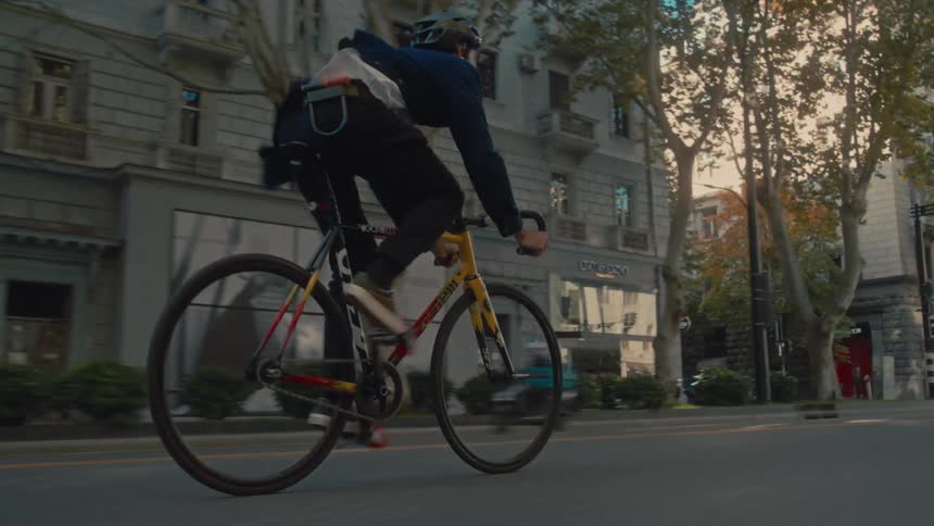 A man pedals a fixedgear bicycle down a city street past buildings and trees while a car drives in the adjacent lane