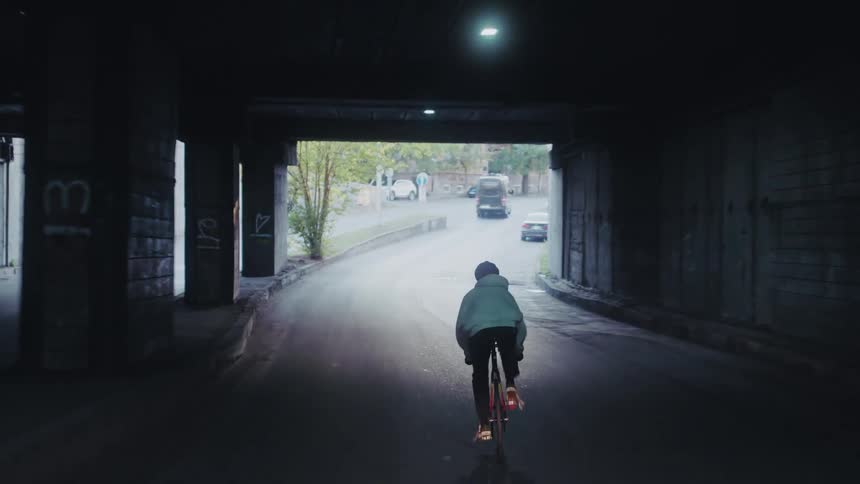 The video shows a cyclist riding a fixedgear bicycle through a dark graffiticovered tunnel or underpass the rider wearing a lightcolored hoodie and dark pants is seen from behind moving towards a patch of light at the far end of the tunnel