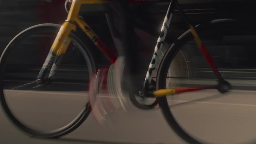 A person is cycling a yellow and red bicycle quickly on a street with the motion of the wheels and legs creating blur while background elements like cars and buildings streak past
