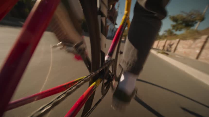 The video is a lowangle closeup shot of the red and yellow wheel spokes and chain of a bicycle in motion the cyclist is wearing dark pants and white socks with dark shoes pedaling down what appears to be a paved road or bridge