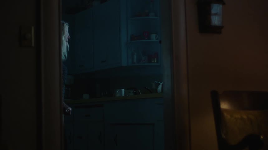A woman stands in a dark kitchen and looks out the window