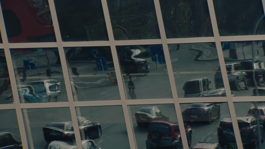A wide view shows the distorted reflection of moving cars and a cyclist on the glass facade of a modern building framed by the white grid of the windows above a parking lot