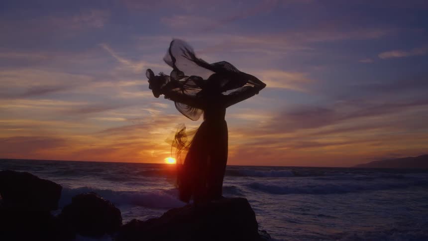 The video is a wide shot of a person silhouetted against a bright sunset over the ocean the figure stands on dark rocks with their arms outstretched holding a piece of sheer fabric that billows around them
