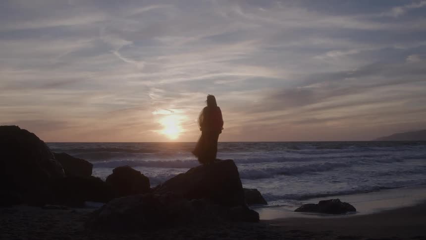 The video shows the silhouette of a person in a flowing dress or robe standing on dark rocks by the ocean the sky is a blend of pale blue and warm sunset colors creating a dramatic mystical atmosphere as waves crash on the shore