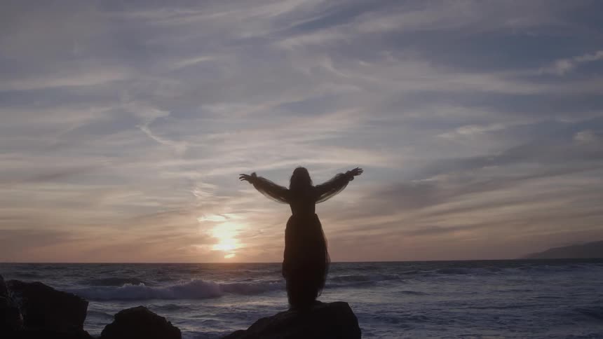 A person stands on a rocky shoreline under a cloudy sky as aggressive waves crash against the rocks sending bursts of sea spray into the air