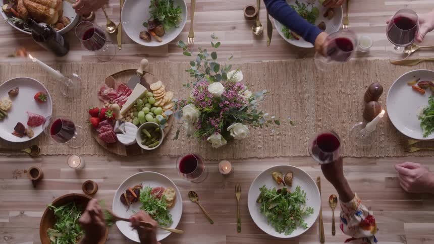 The video is an overhead shot of a dinner party table set with a variety of food including a large roast salad and a charcuterie board hands are visible reaching for the dishes and clinking glasses of red wine