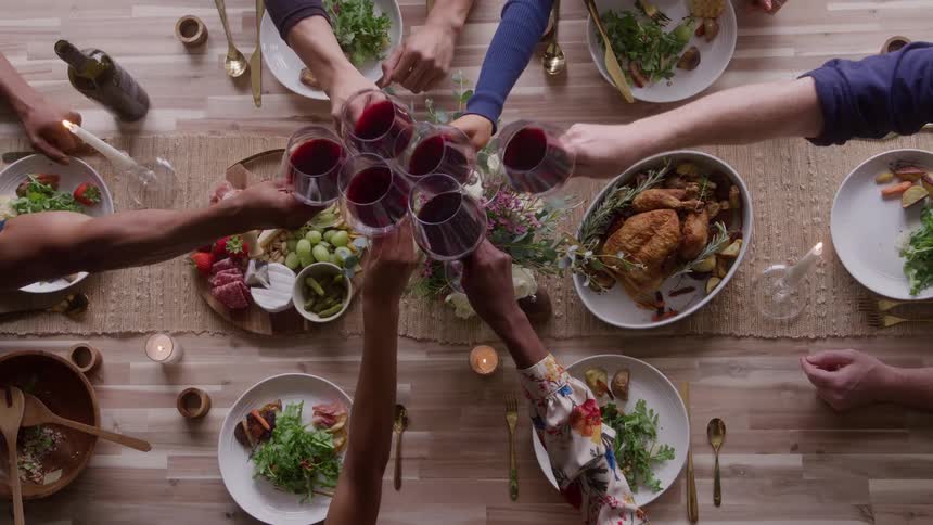 The video is an overhead shot of a dinner party table set with food wine and candles multiple peoples hands are visible reaching for food and clinking glasses of red wine