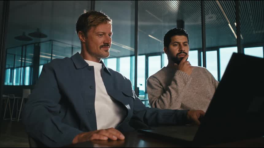 Two men viewing media on a laptop computer while sitting together at a table in an office