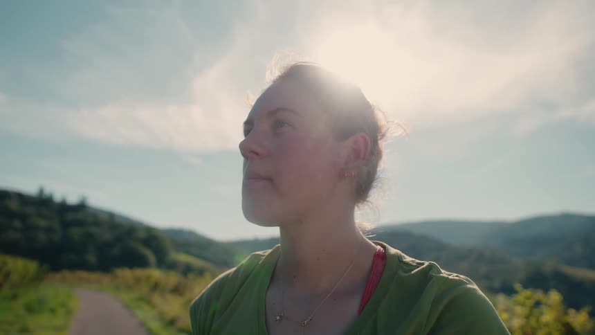A close profile view of a smiling woman with her hair in a bun wearing a green shirt shows her eating a piece of fruit outdoors in a sunny vineyard with rolling green hills in the background