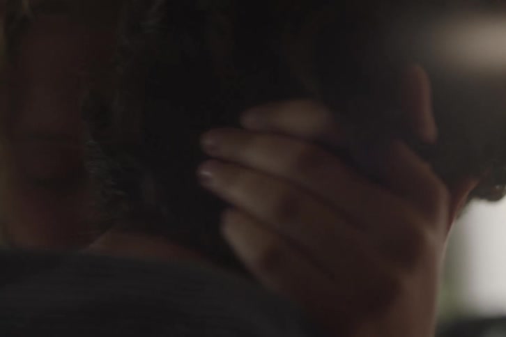 An extremely closeup shot focuses on the heads and upper backs of a man and a woman with one persons face pressed near the others neck and ear engaging in a close embrace in a dimly lit indistinct space