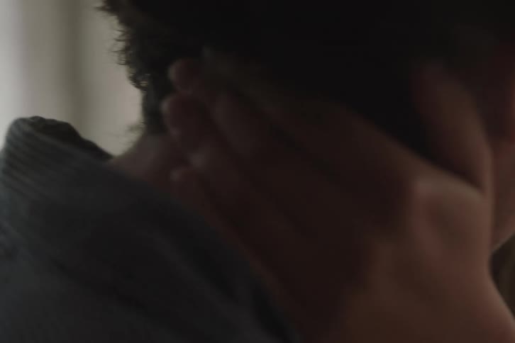 A close dark intimate shot shows a womans hands gently caressing the neck and hair of a man wearing a striped collared shirt as they embrace in soft lighting