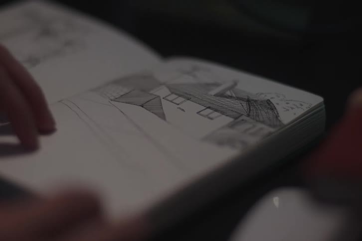 A close view shows a hand slowly turning the pages of an open notebook in a dimly lit setting revealing both pages filled with handwritten notes and pages with pencil sketches