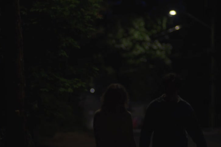 A medium dark shot shows a couple walking slowly hand in hand on a quiet street at night with the woman wearing a yellow top and their figures fading into the darkness as streetlights blur