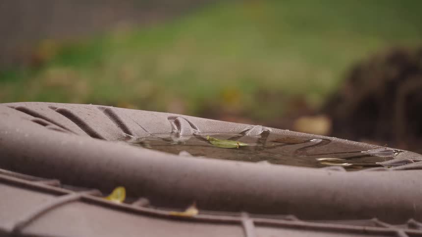 A low close view shows rain drops creating ripples in a small puddle of water collected on the brown textured plastic lid of an outdoor barrel