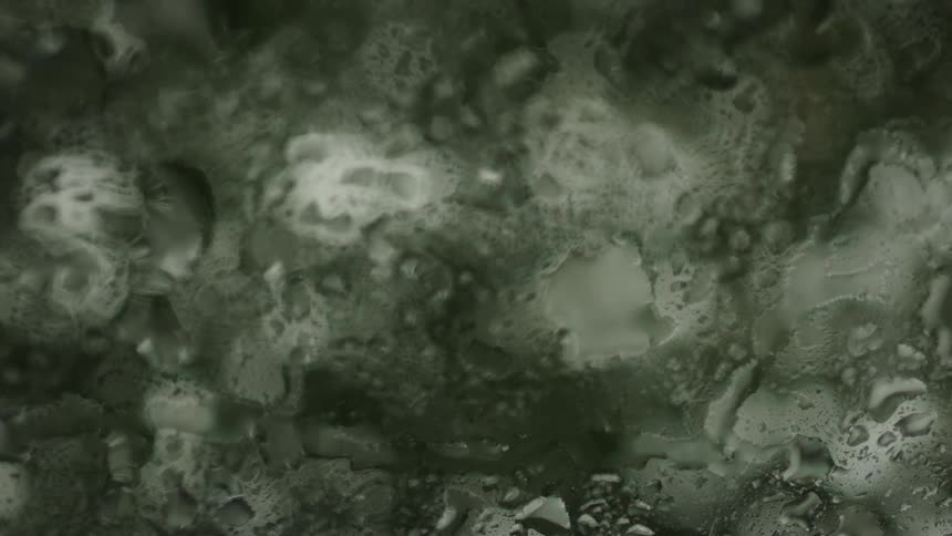 A very close abstract view of a window shows heavy rain creating large water droplets and flowing streams across the glass pane with a blurred dark green background