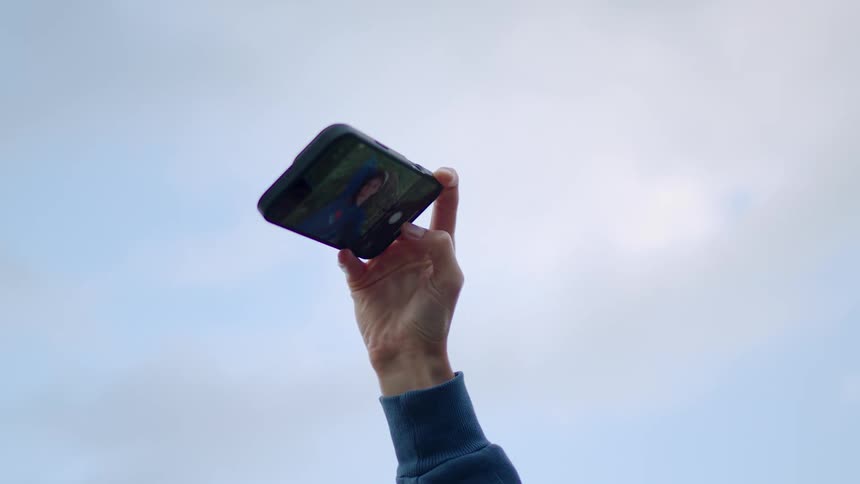 A hand in a blue sleeve holds a smartphone high against a pale sky with a selfie visible on the screen