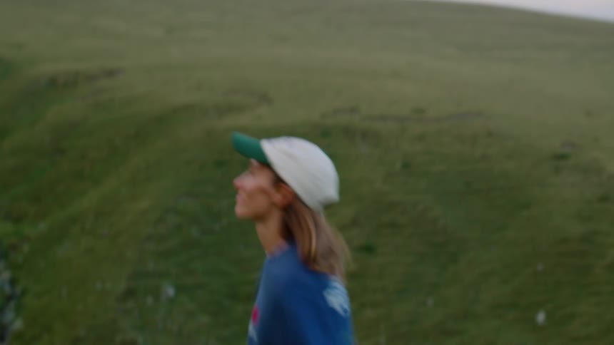 Woman walking on a green hillside wearing a blue sweatshirt and light cap