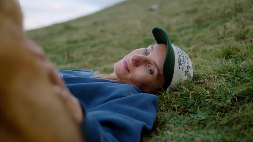 A woman wearing a cap lies in the grass looking up lovingly at her dog