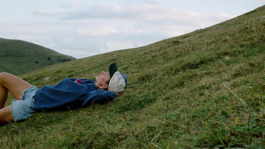 A female in a blue sweatshirt and cap lies back on a grassy hillside with hands behind the head gazing up in quiet relaxation
