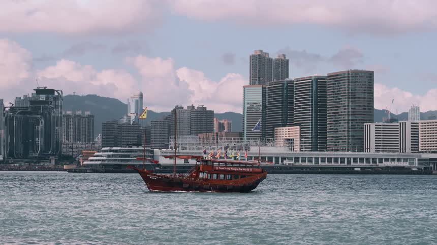 An orange water vessel with multiple flags moving across a body of water beside a metropolitan city during daytime