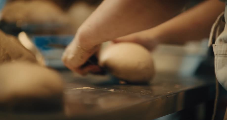 A baker rolling and shaping dough into a round ball on a stainless steal work surface