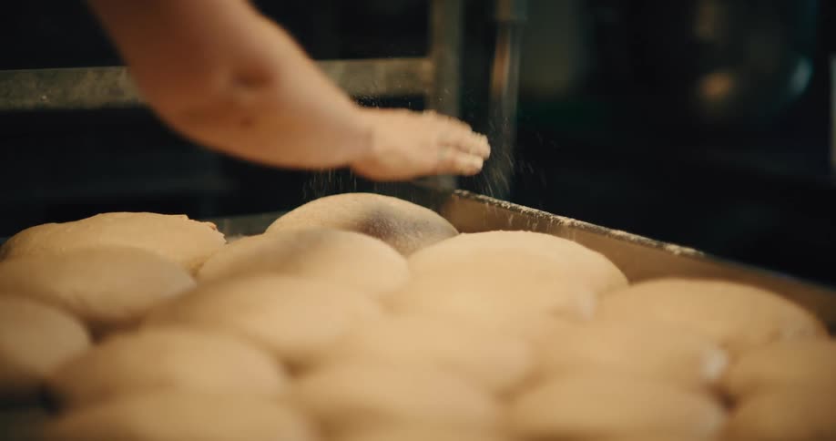 Baker sprinkling flour over portions of dough arranged on a tray
