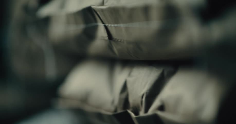 A blurry extreme close view shows the stitched seams of a row of brown paper bags that are rapidly moving by on a conveyor belt indicating industrial packaging in a factory setting