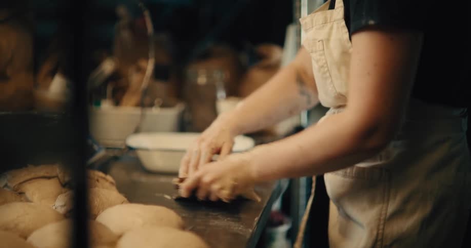 A baker wearing glasses a dark shirt and a white apron is seen from the chest down carefully shaping loaves of light colored bread dough by hand on a counter in a dimly lit bakery kitchen
