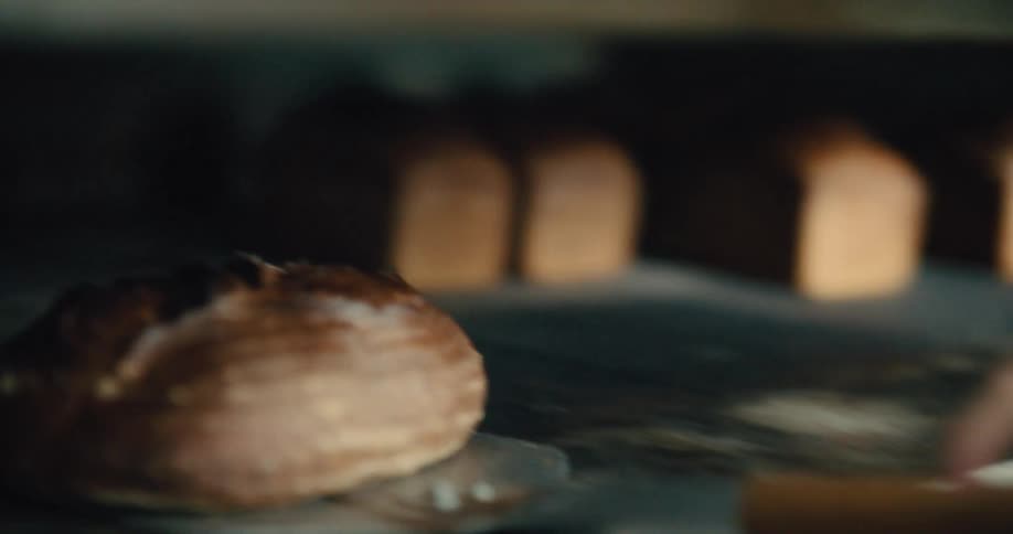 A baker wearing a white apron uses a baker's paddle to remove dark sourdough bread from a hot oven