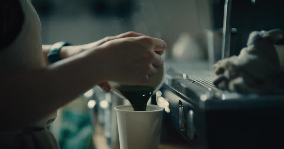 A persons hands holding a white ceramic cup over a tall disposable cup and pouring a dark green liquid that emits a small amount of steam into the disposable cup