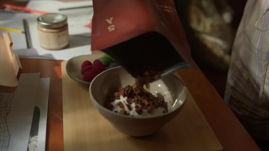 Person adding granola to a bowl of yogurt on a wooden table