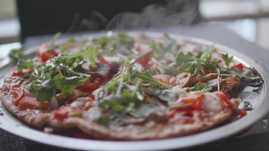 The video is a closeup shot of a freshly baked steaming pizza or flatbread topped with colorful ingredients including sliced red onions tomatoes mushrooms and a generous layer of fresh arugula the food is resting on a circular metal pan and